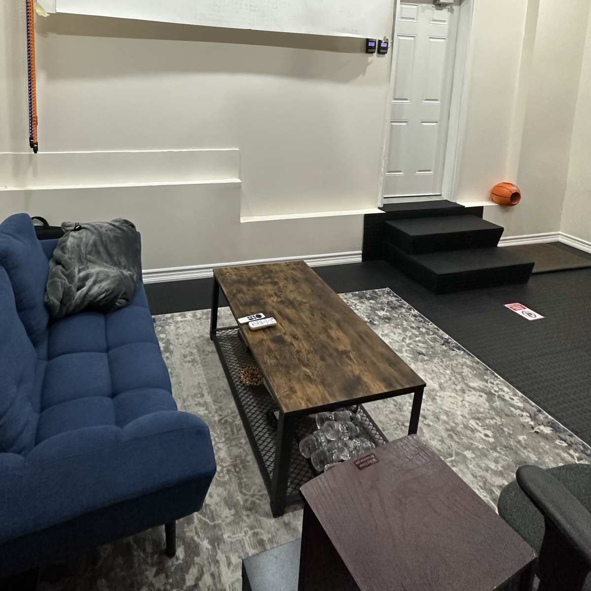 A blue couch and wooden coffee table are positioned in a room with light-colored walls and a door leading outside, along with a rug and steps in the background.
