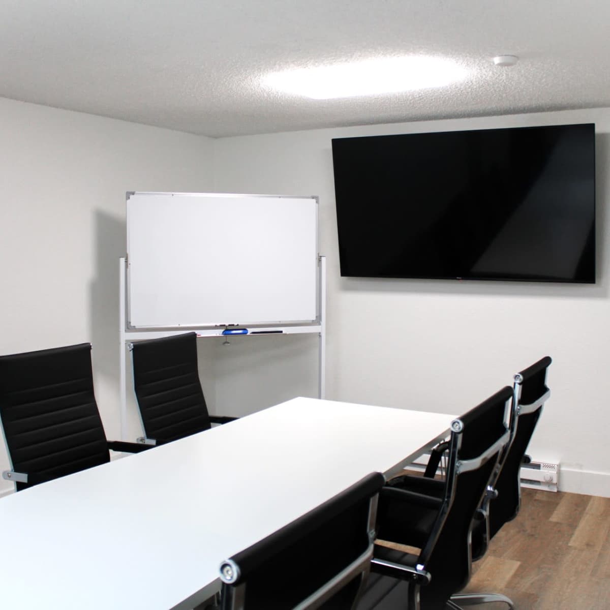 A modern conference room features a long white table, six black chairs, a whiteboard on a stand, and a wall-mounted TV.