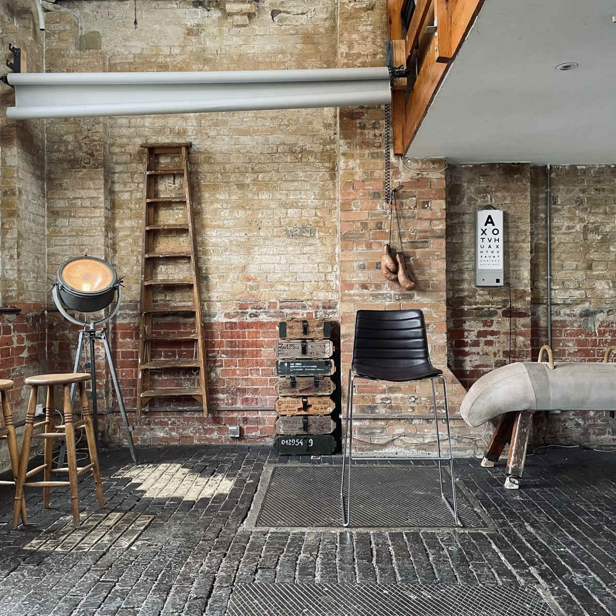 An industrial-style room with exposed brick walls, wooden ladders, a spotlight, a black chair, two wooden stools, and a vintage pommel horse.