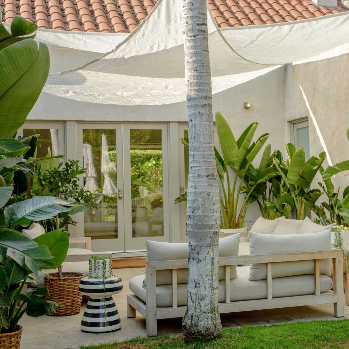 A shaded outdoor seating area is lined with potted plants and features a wooden couch with cushions.