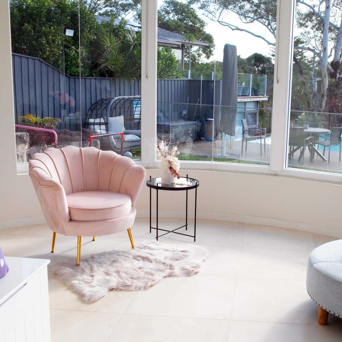 A pink chair and a round ottoman are positioned next to a small table in a room surrounded by large windows overlooking a garden with outdoor seating.