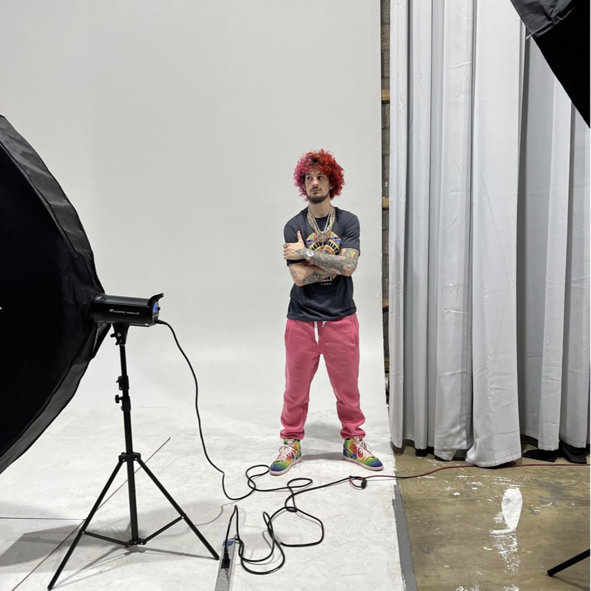 A person with red hair stands in a studio with large photography lights on either side and a plain backdrop behind them.