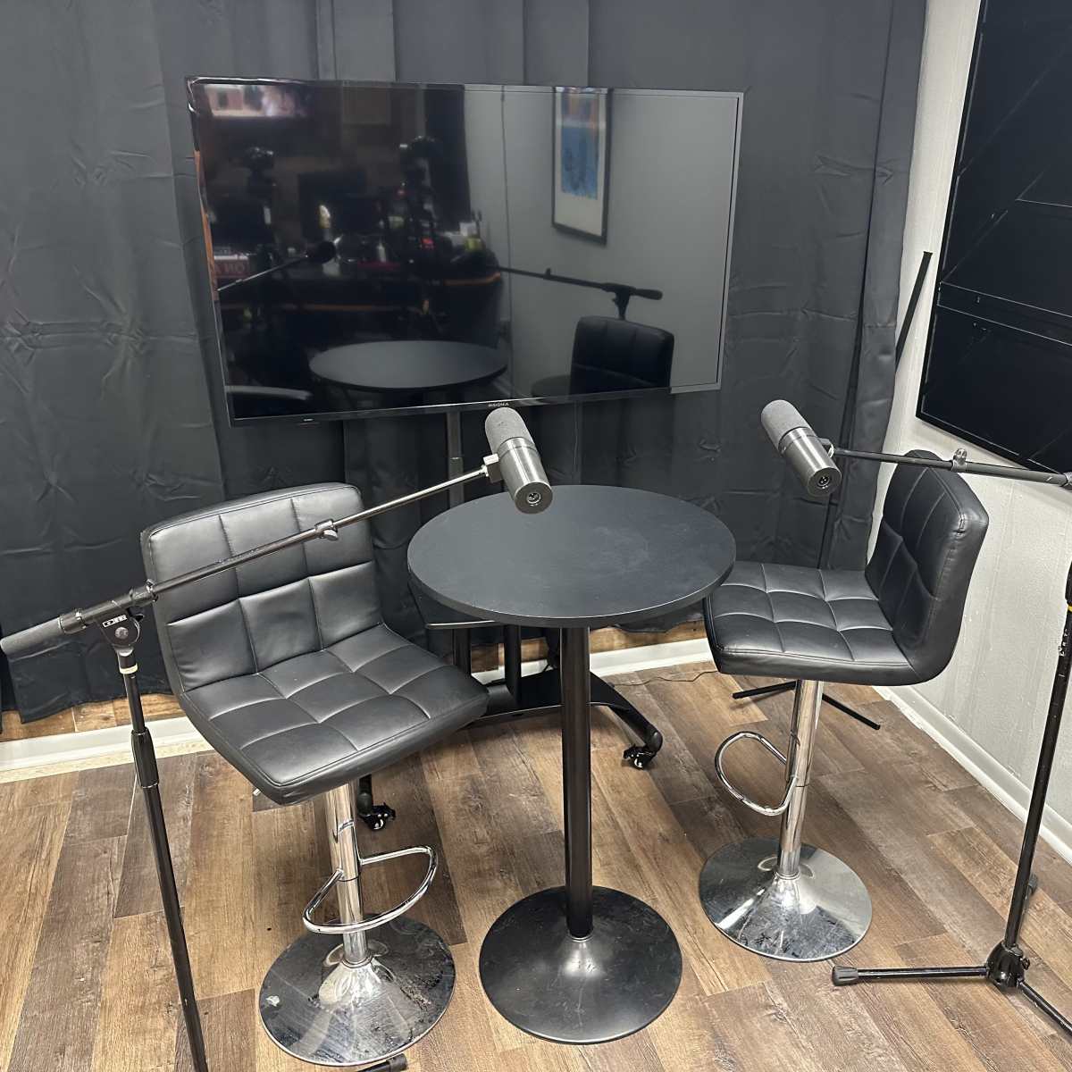 A small recording studio setup featuring two black chairs, a round table, and two microphones on stands, with a television mounted on the wall in the background.