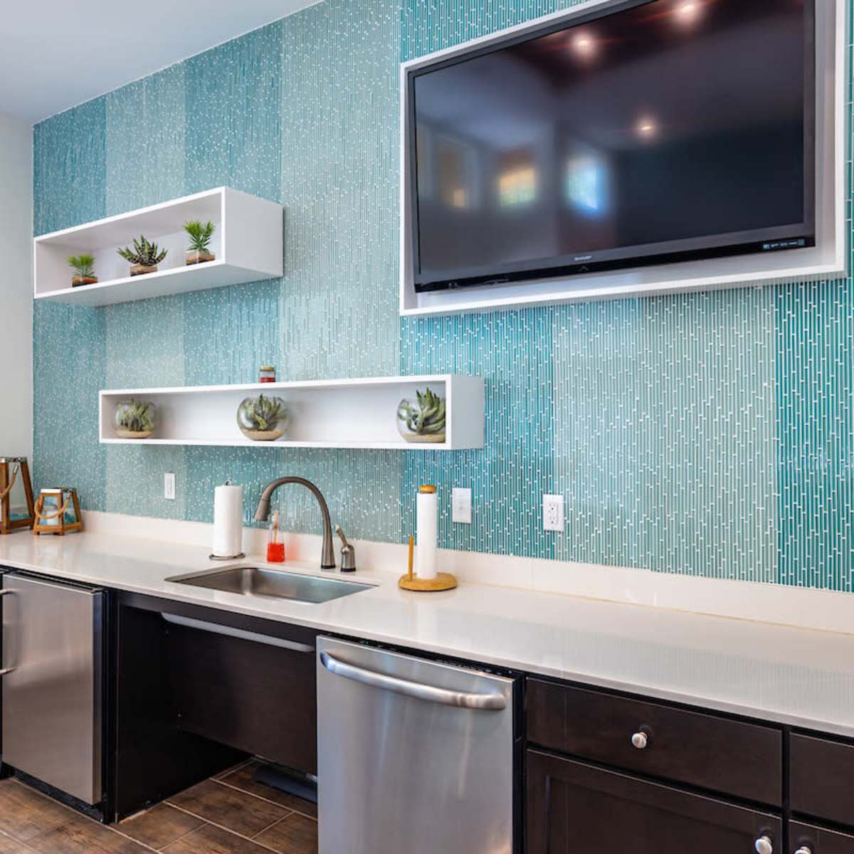 A modern kitchen with a turquoise and white striped backsplash, a stainless steel sink, a television mounted on the wall, and shelves displaying small plants.