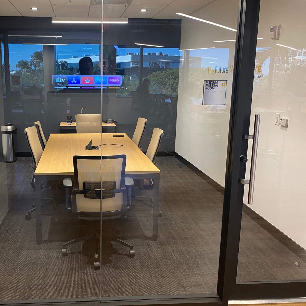 A modern conference room with a table and six chairs, viewed through a glass wall, featuring a large screen displaying a presentation.