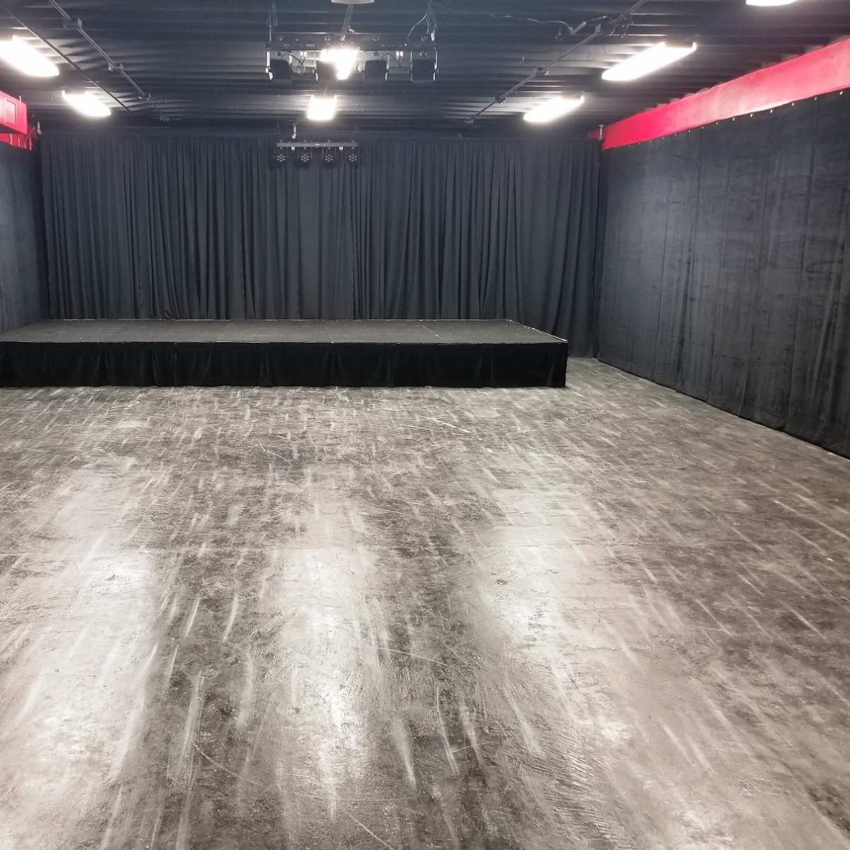 An empty performance space with a raised stage and black curtains in a dimly lit room.