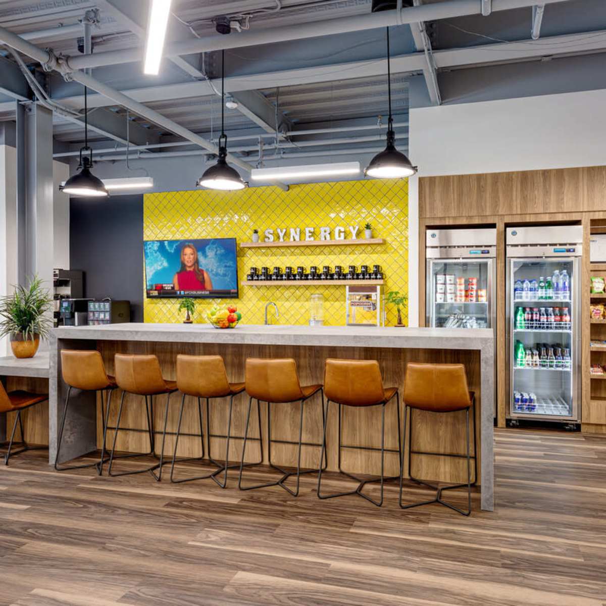 A modern break room with a long bar counter, orange stools, a vending machine, and a wall-mounted television displaying a video.