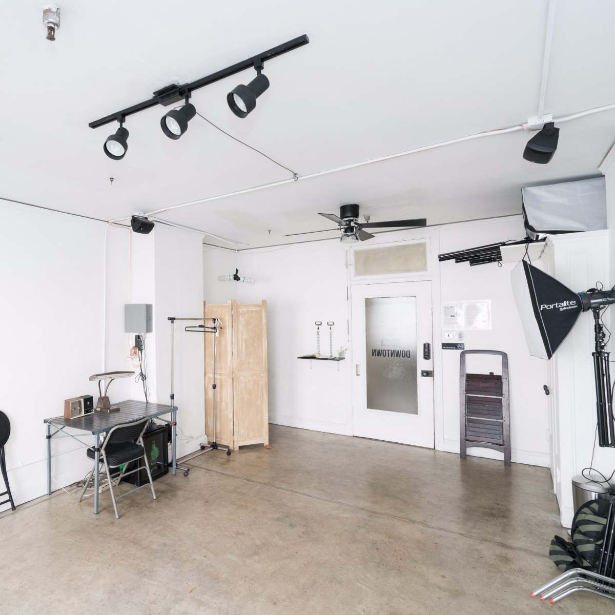 A minimalistic room features a desk with chairs, a wooden wardrobe, and lighting equipment against white walls and a concrete floor.