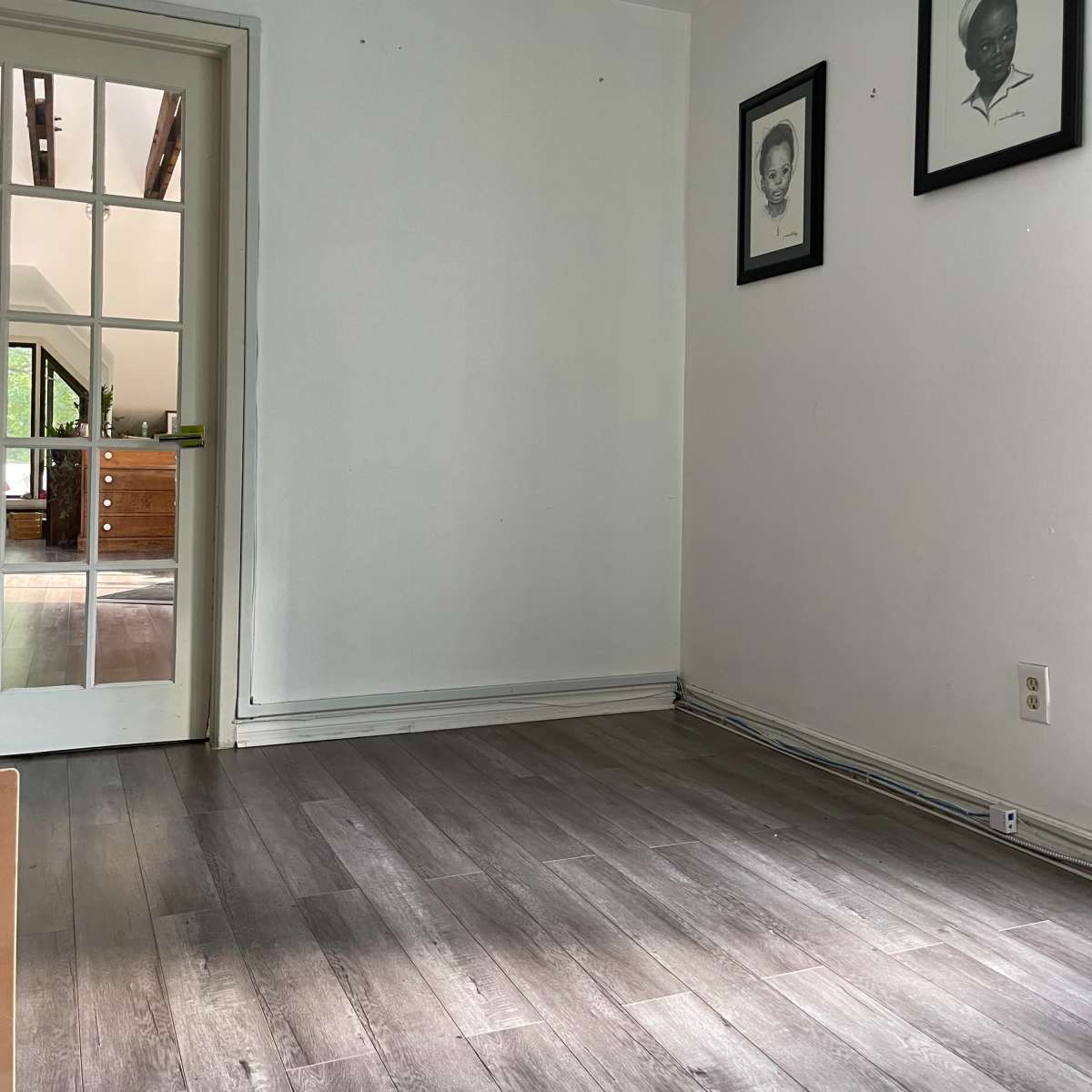 A sparse room with light-colored walls, a wooden floor, and two framed artworks hanging on the wall.