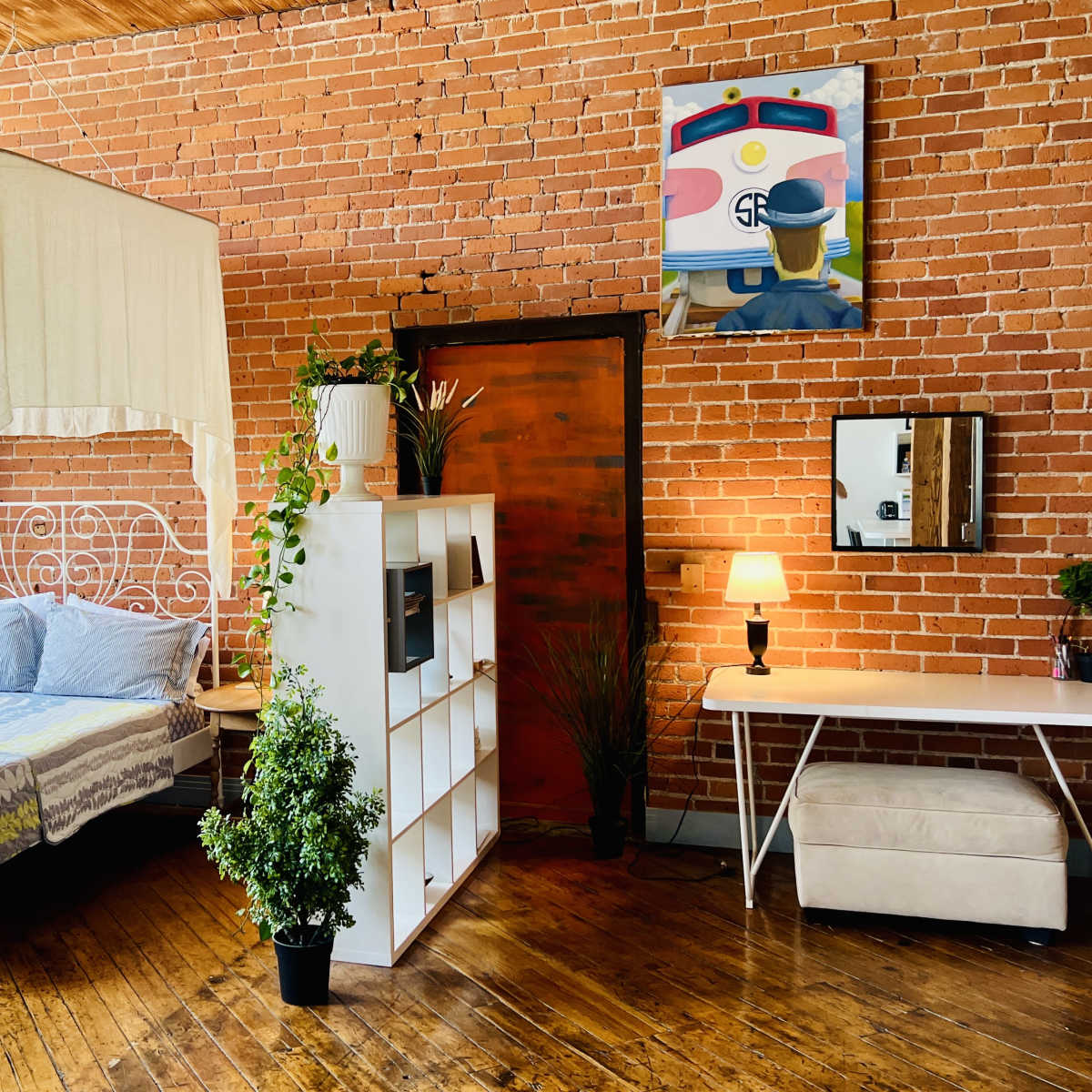 A bedroom with a white canopy bed, a bookshelf, a desk, and brick walls, featuring a painting of a train.