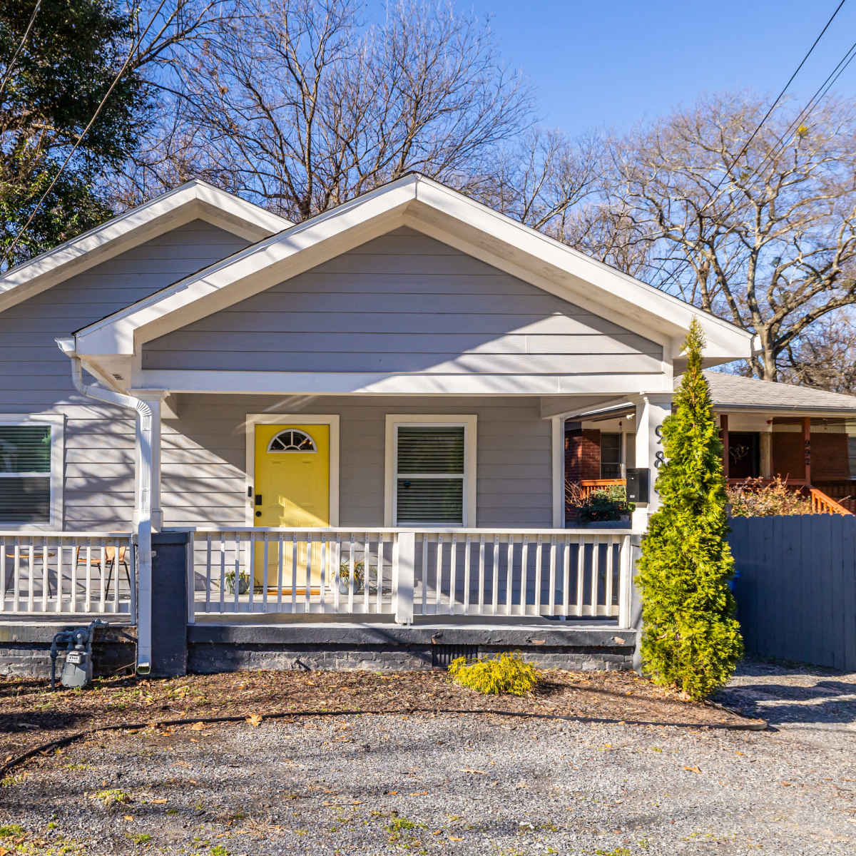 A single-story gray house with a yellow front door and a porch is surrounded by a gravel driveway and a low stone wall.