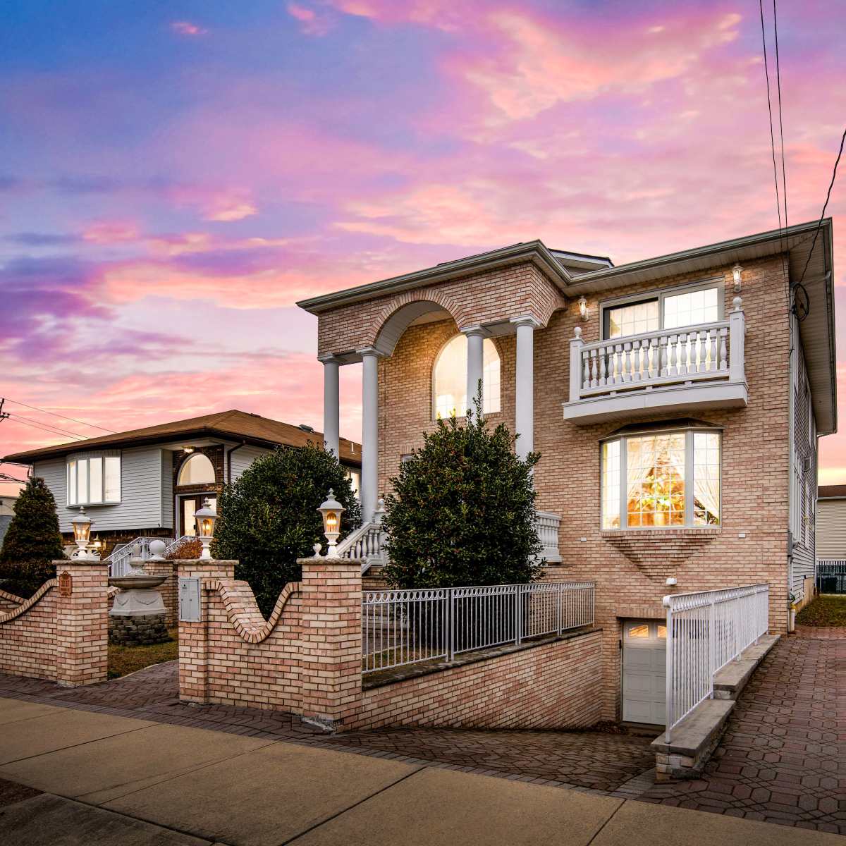 A two-story brick house with a balcony and landscaped front yard is illuminated against a vibrant sunset sky.