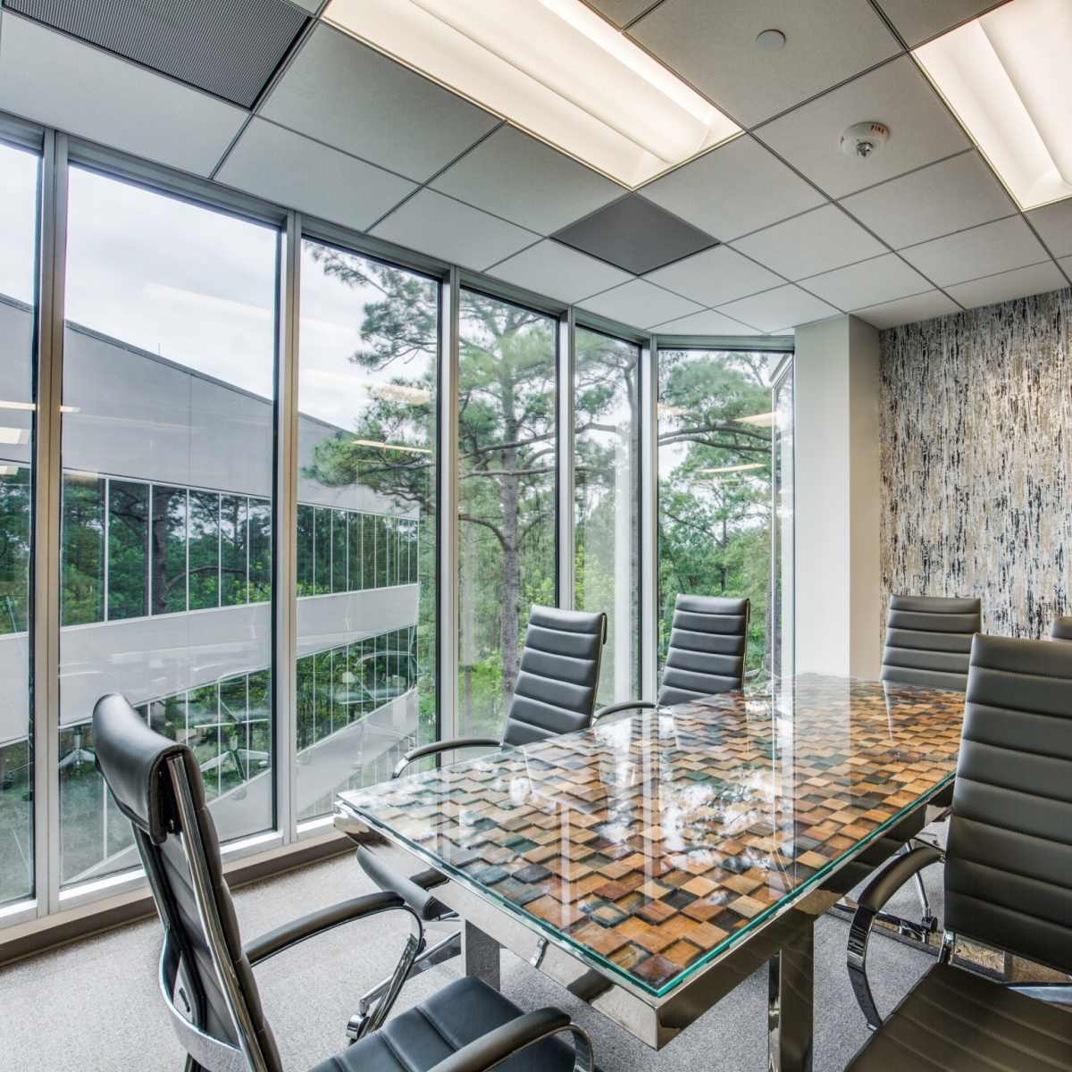A conference room features a glass-topped table surrounded by black leather chairs, with large windows displaying a view of trees.