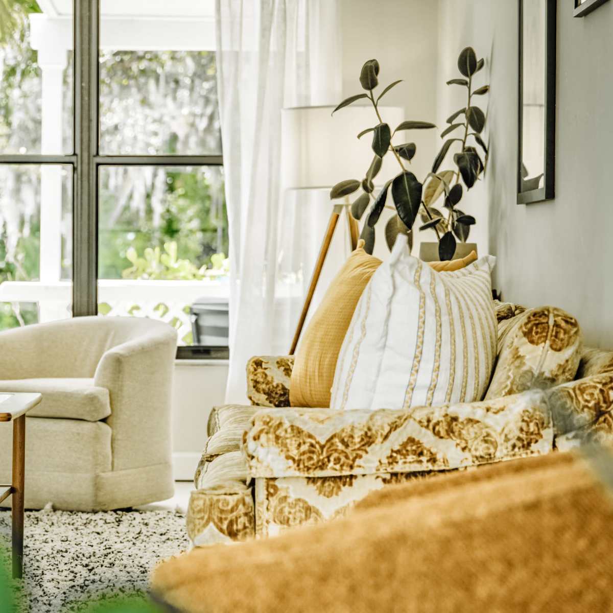 A beige sofa and armchair are positioned in a brightly lit living room with large windows and green plants visible outside.
