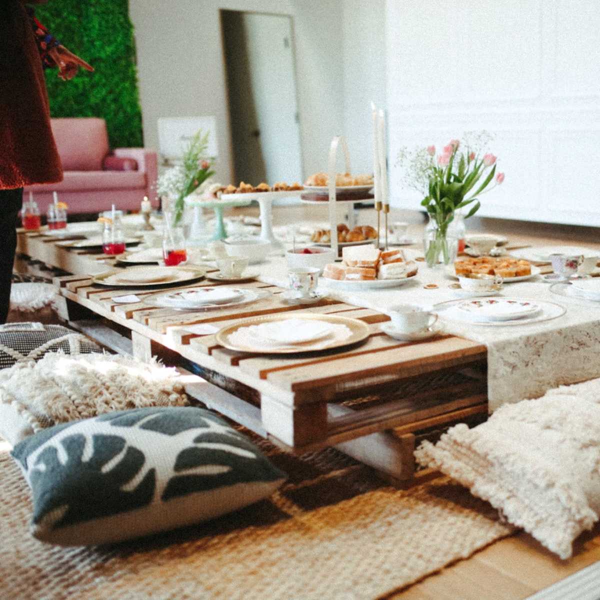 A low wooden table is set with tea cups and various snacks, surrounded by cushions on a woven rug in an indoor space.