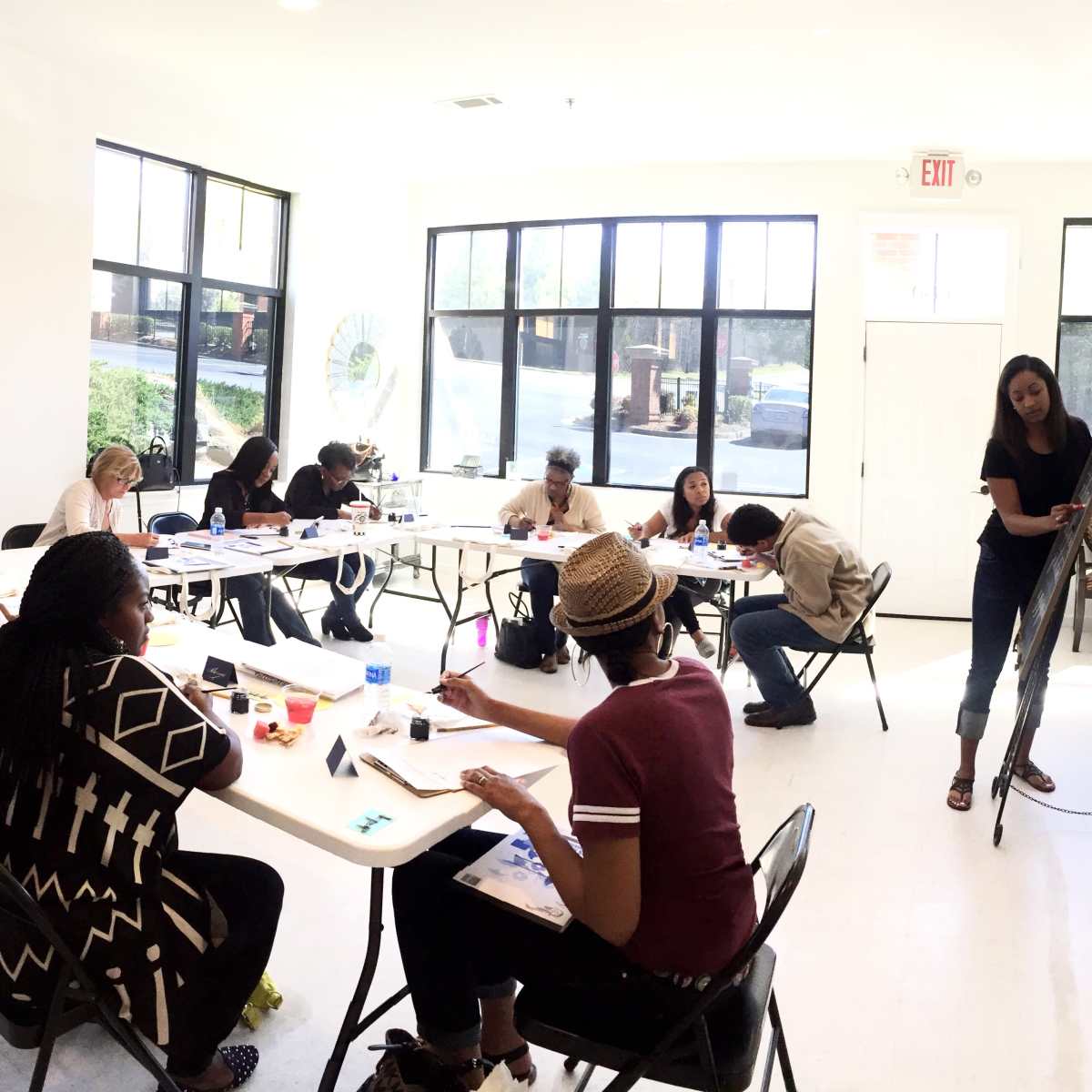 A group of people participates in an art class, sitting at tables with art supplies while one instructor demonstrates techniques.