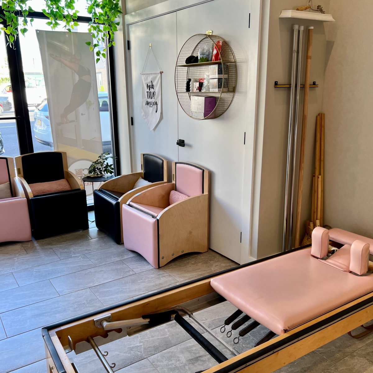 A wellness studio with a pink reformer machine, various seating options, and a wall decorated with greenery and shelves.