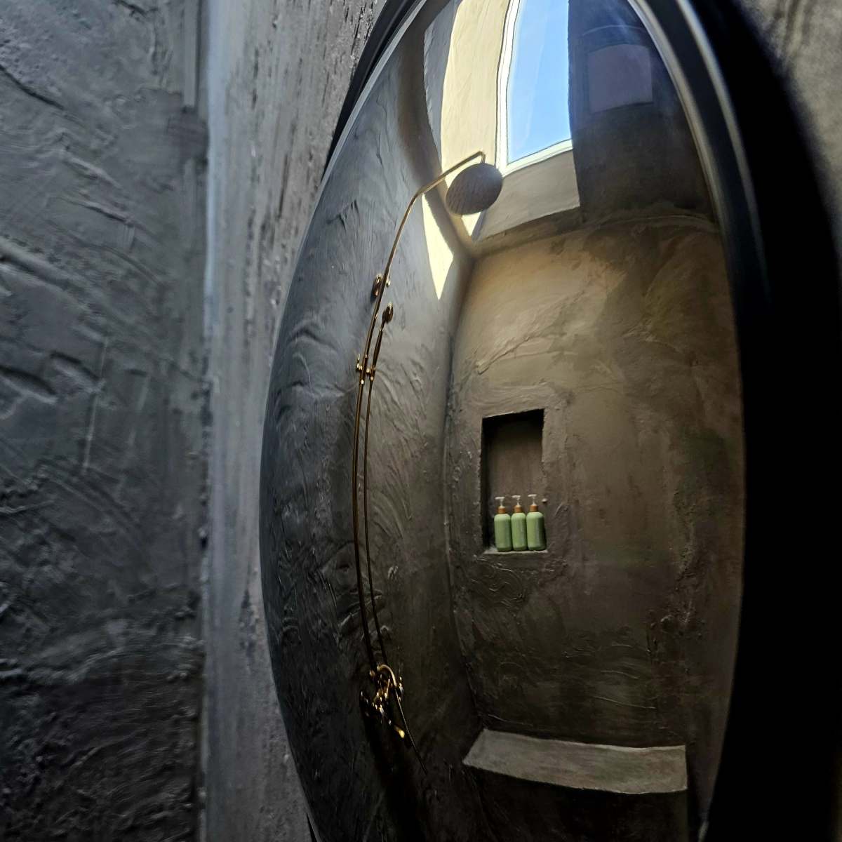 A round mirror reflects a dimly lit corner of a textured gray bathroom with a shelf holding three green bottles.