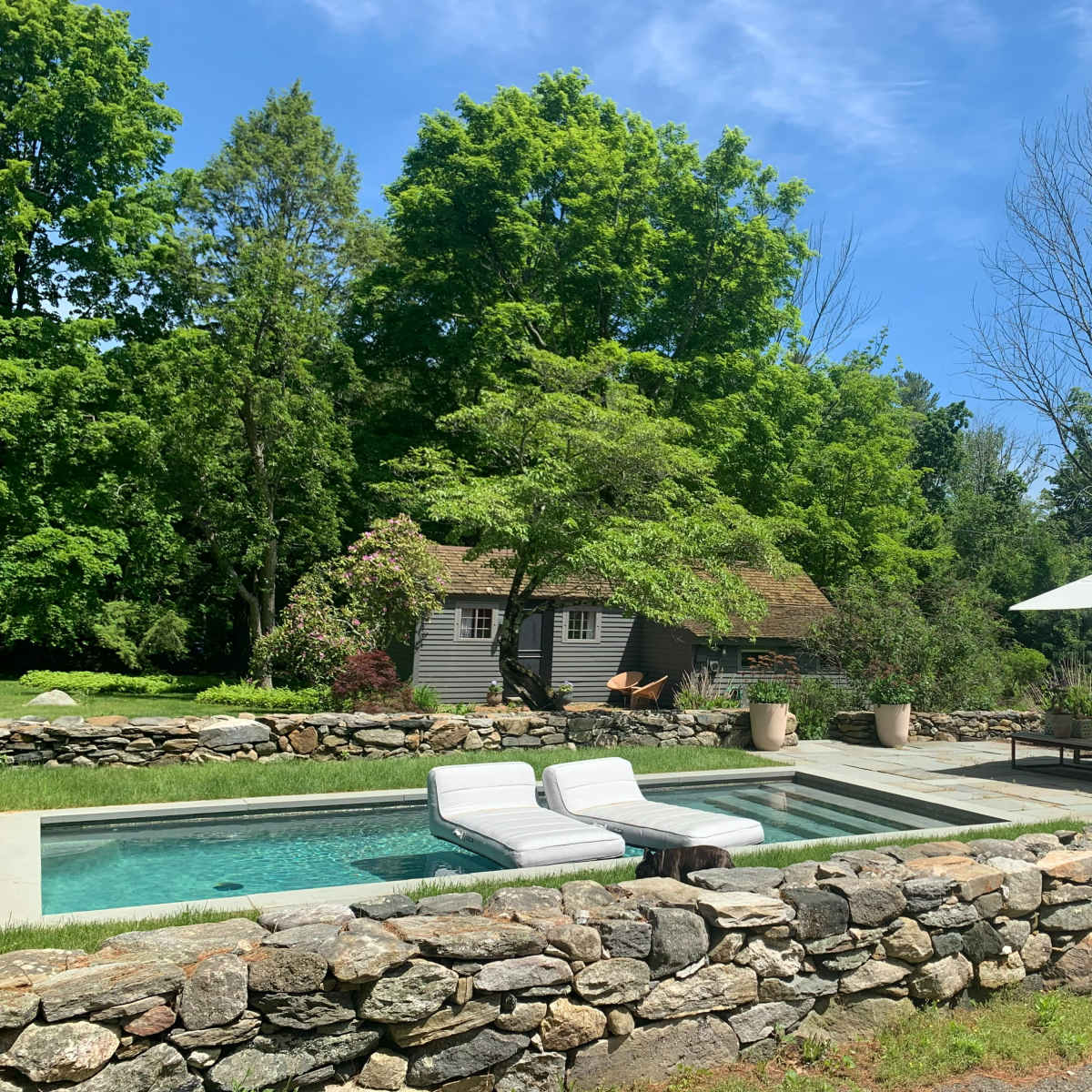 A rectangular pool with lounge chairs is surrounded by a stone wall and greenery, with a shaded seating area and a rustic building in the background.