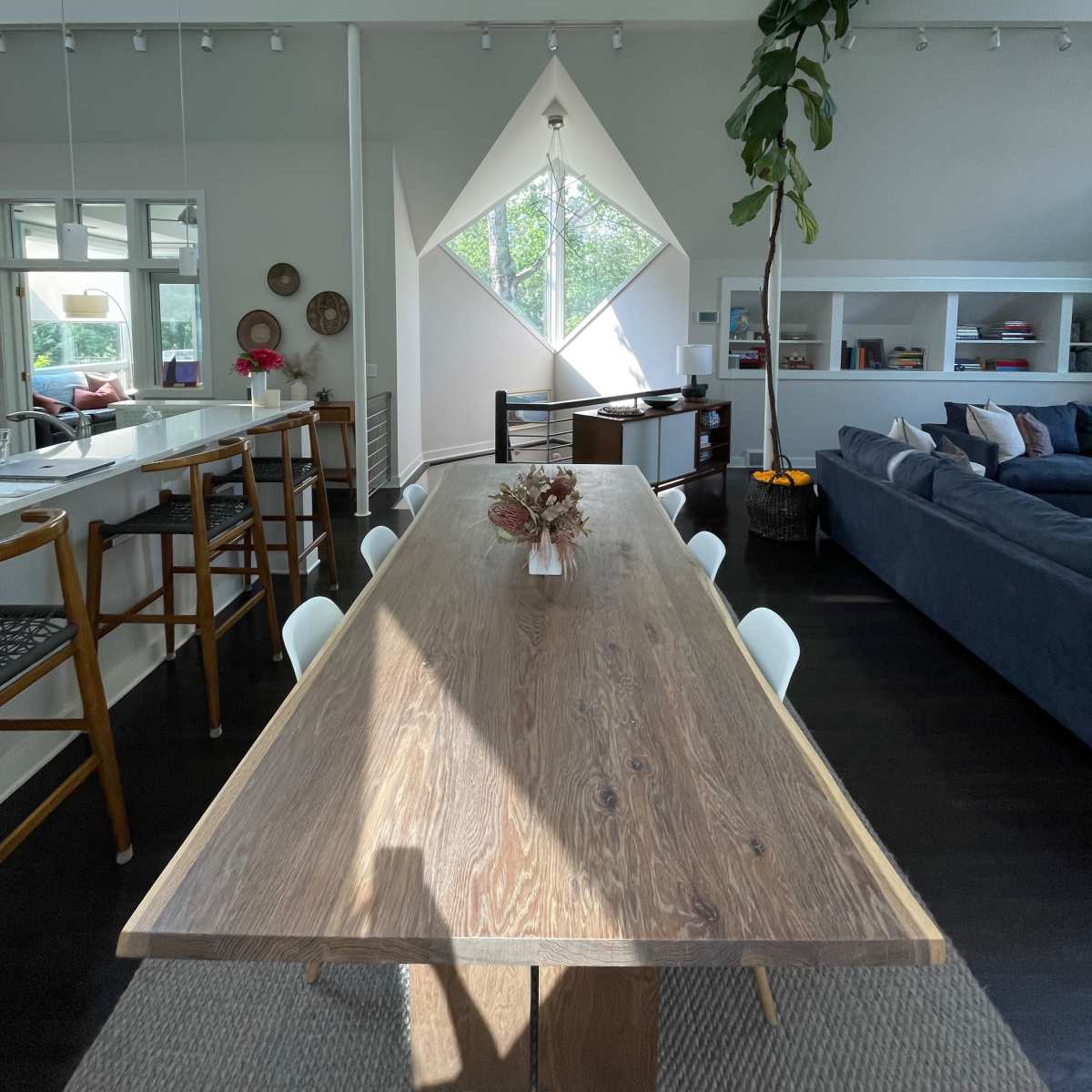 A modern interior features a long wooden dining table, a large diamond-shaped window, and a living area with a blue sofa and greenery.