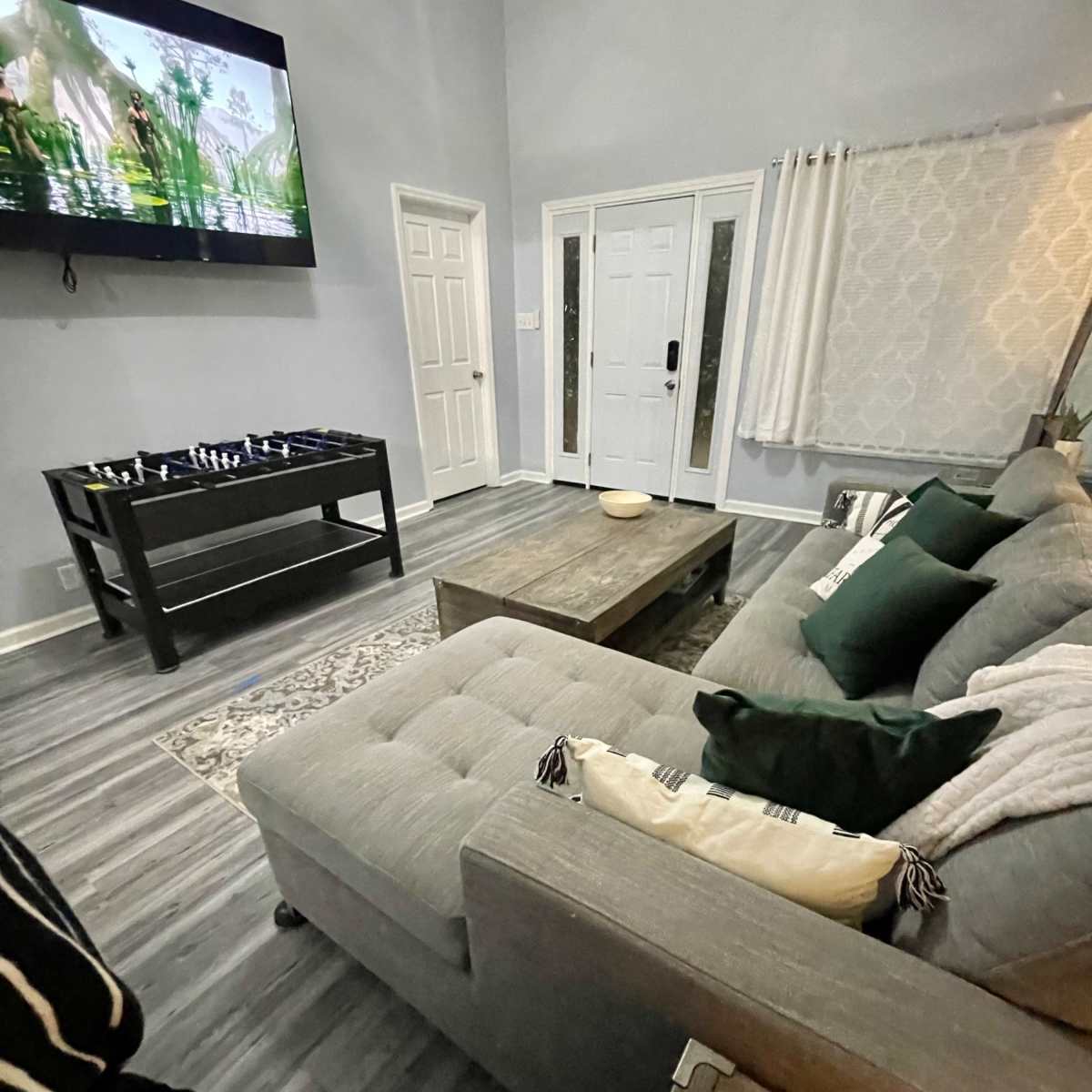 A living room featuring a grey sofa, a wooden coffee table, a television mounted on the wall, and a front door visible in the background.