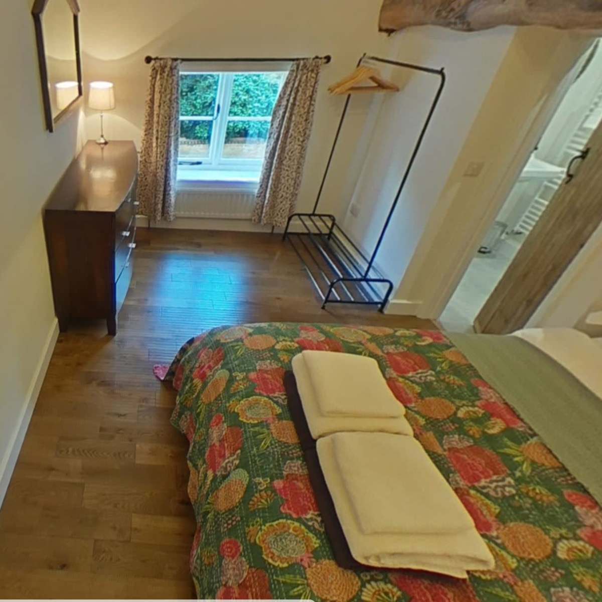 A bedroom with a colorful bedspread, a wooden dresser, and a window revealing greenery outside.