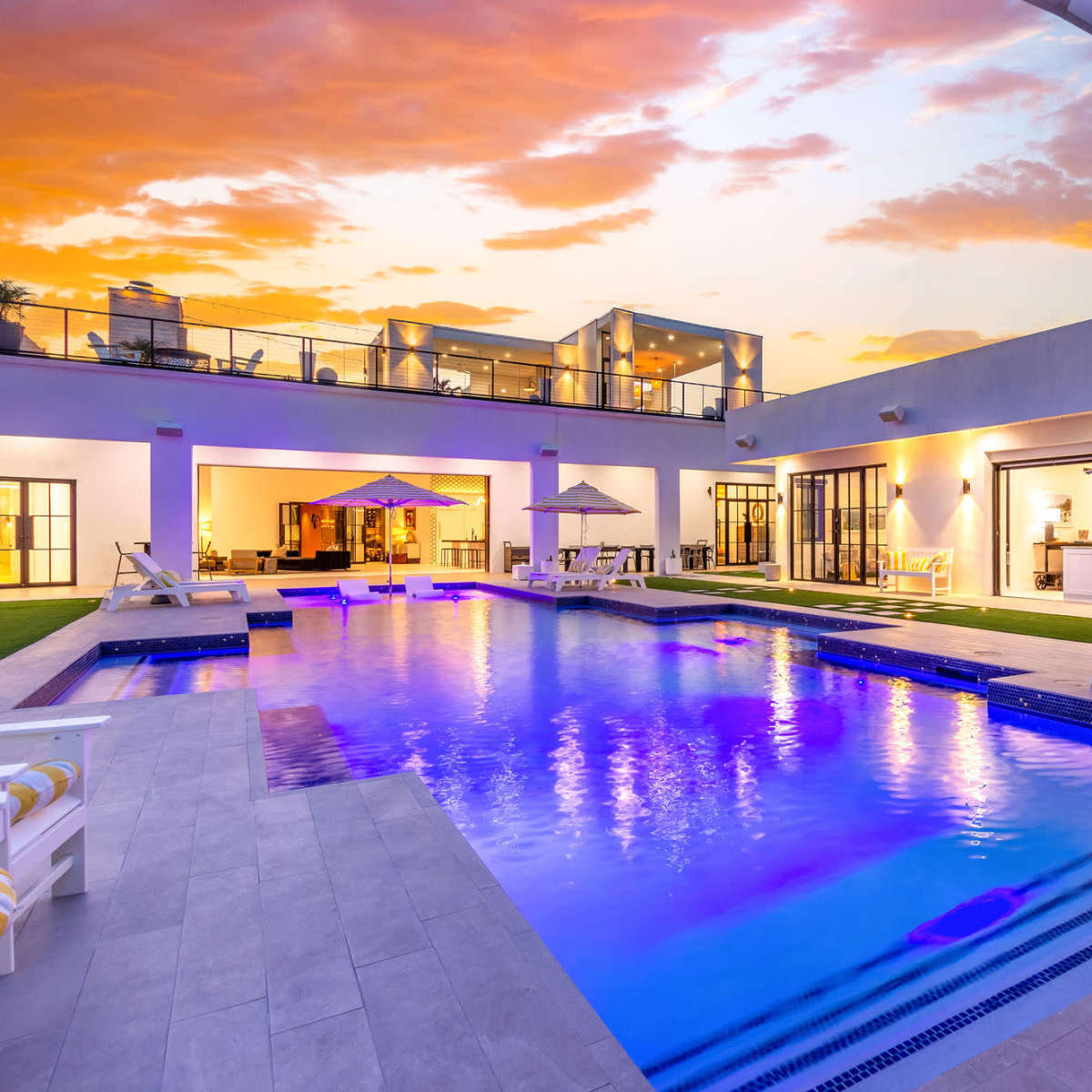A modern outdoor pool area features lounge chairs, umbrellas, and a backdrop of a sunset sky.