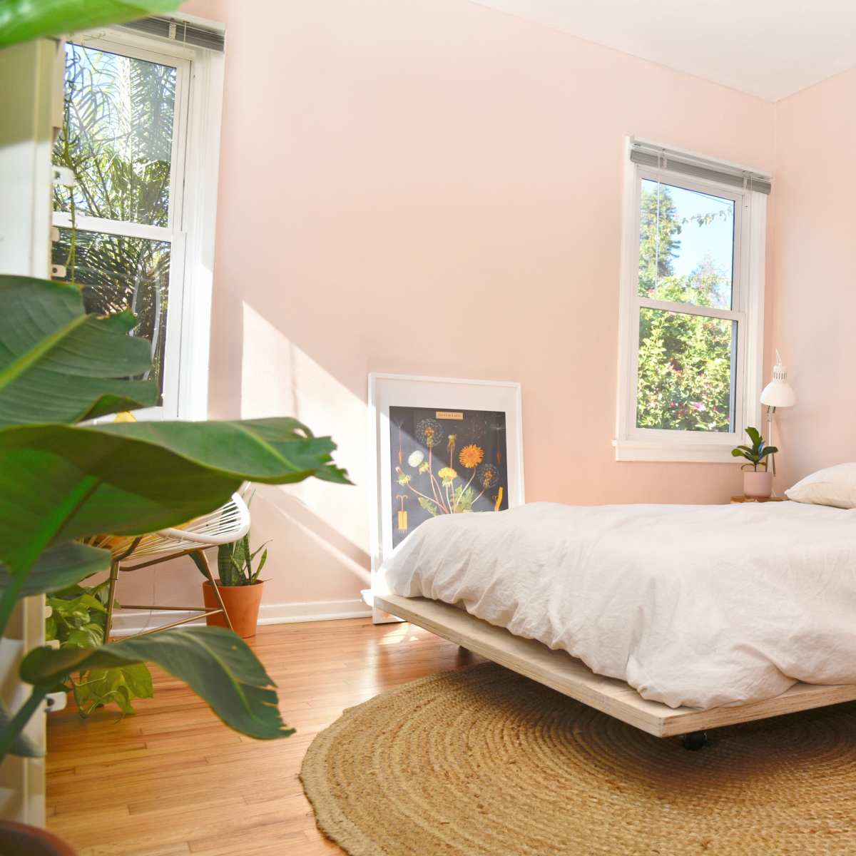 A minimalistic bedroom with a light-colored bed, a round area rug, and plants beside large windows.