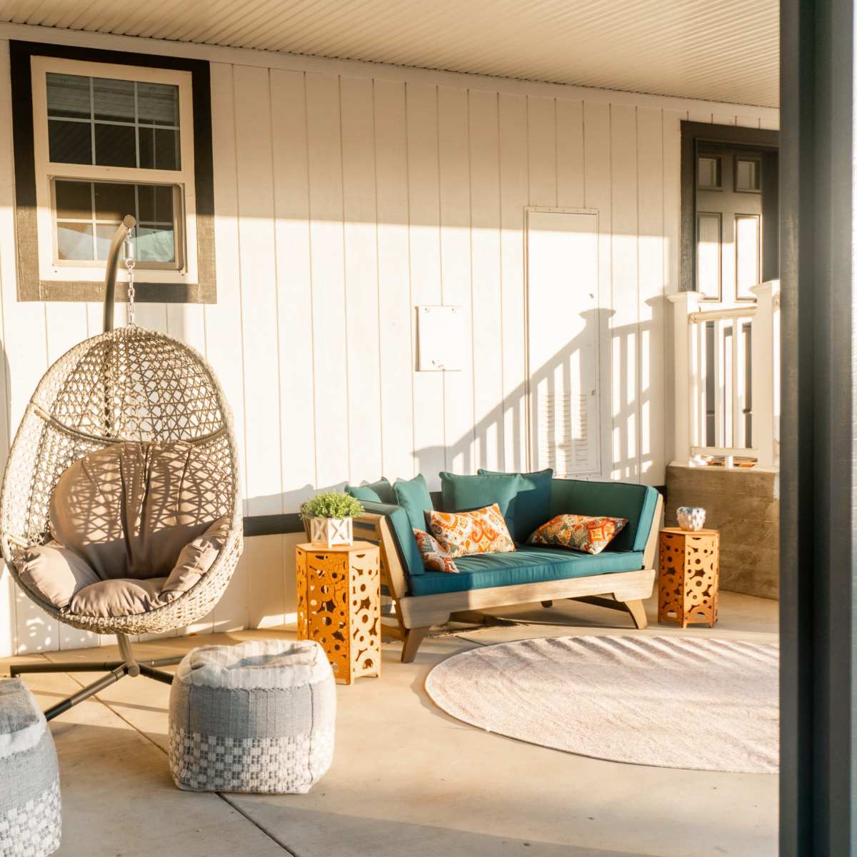 A stylish porch area with two hanging chairs, a teal couch, and decorative pillows, accompanied by light-colored poufs and a round rug.