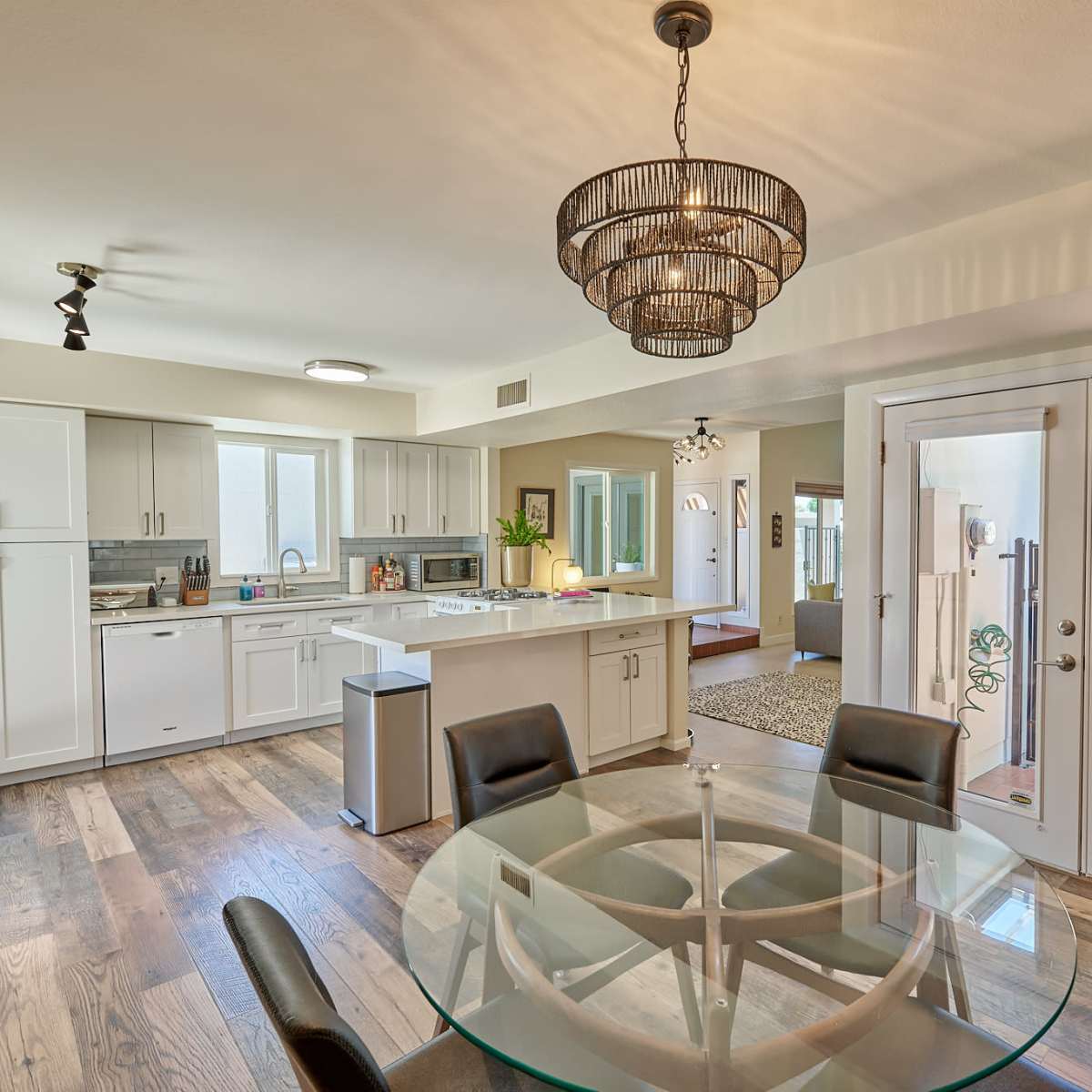 A modern kitchen and dining area featuring a round glass table surrounded by black chairs, with bright cabinetry and an open layout leading to an exterior door.
