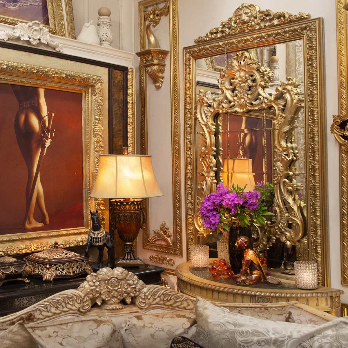 An ornate room featuring gilded mirrors, a framed painting of legs, a lamp, and a vase of purple flowers on a decorative table.
