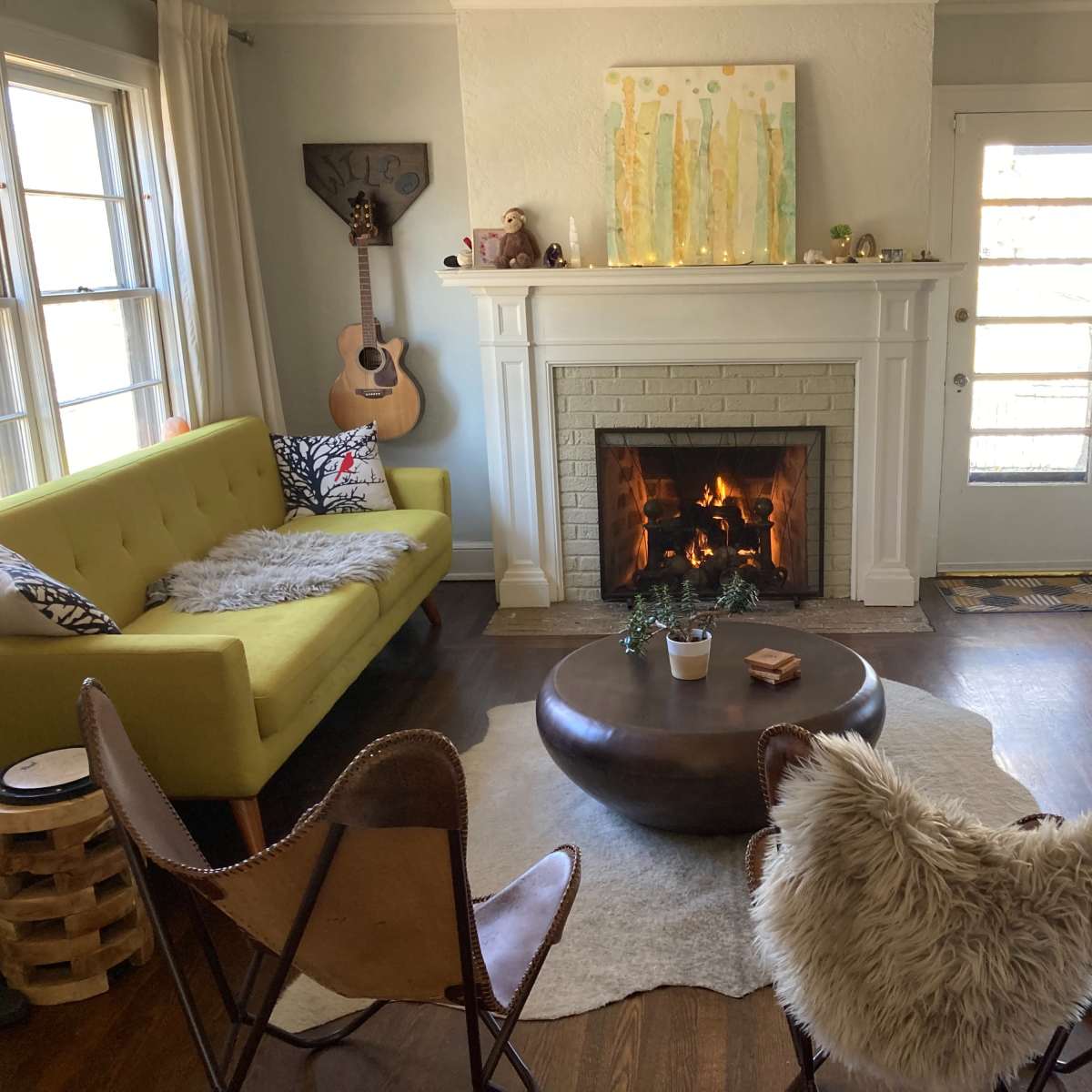 The living room features a yellow sofa, two leather chairs with fur throws, a round coffee table, and a fireplace with a fire burning in it.