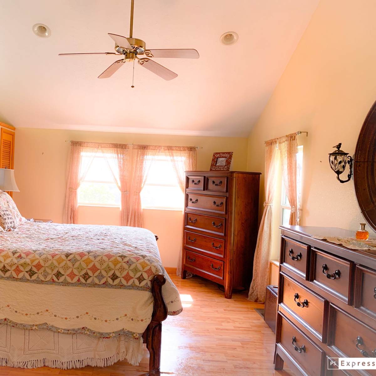 A well-furnished bedroom featuring a large bed, wooden dressers, and windows with light-colored curtains.