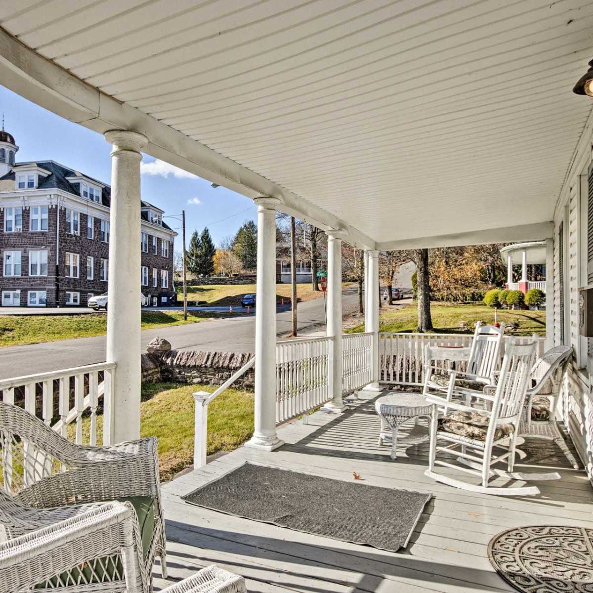 A porch with white rocking chairs, overlooking a street and a large building in the background.
