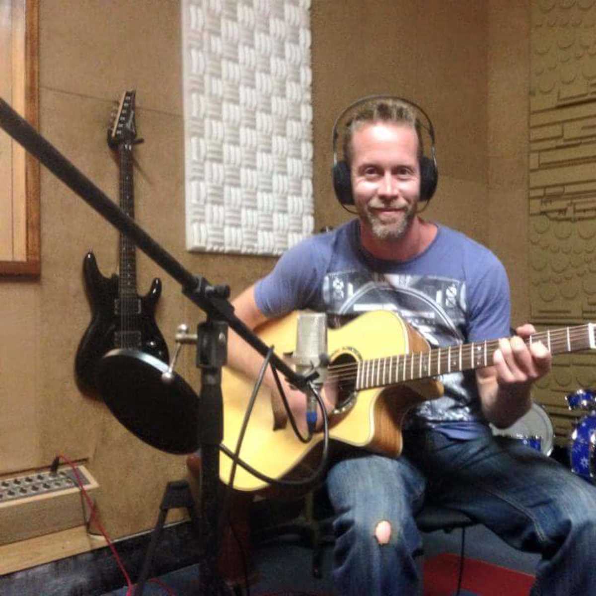A man with headphones plays an acoustic guitar in a sound recording studio.