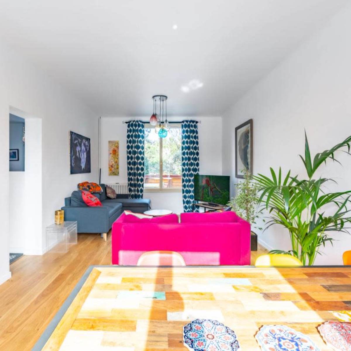 A bright living space with a dining table in the foreground, a gray sofa, and colorful decor, including curtains and cushions.