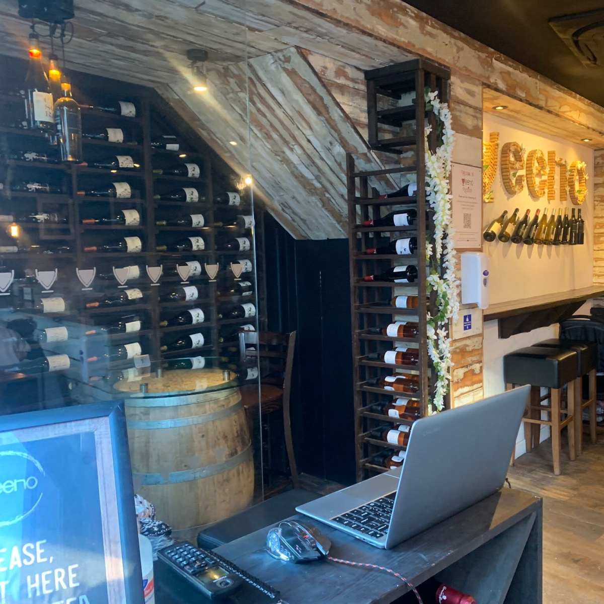 A wine bar interior with a wooden wall, wine bottles neatly arranged on shelves, and a laptop on a black counter.