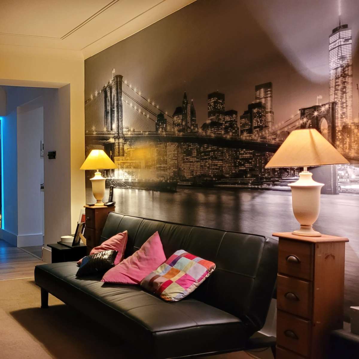 A living room features a black sofa with colorful cushions, a side table with a lamp, and a dining area illuminated by blue and yellow lights in the background.