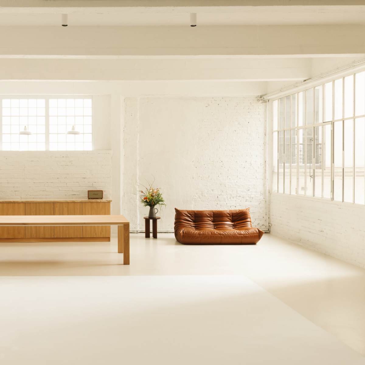 A spacious, minimalist interior with a wooden kitchen, a large dining table, and a brown sofa near a window with natural light.