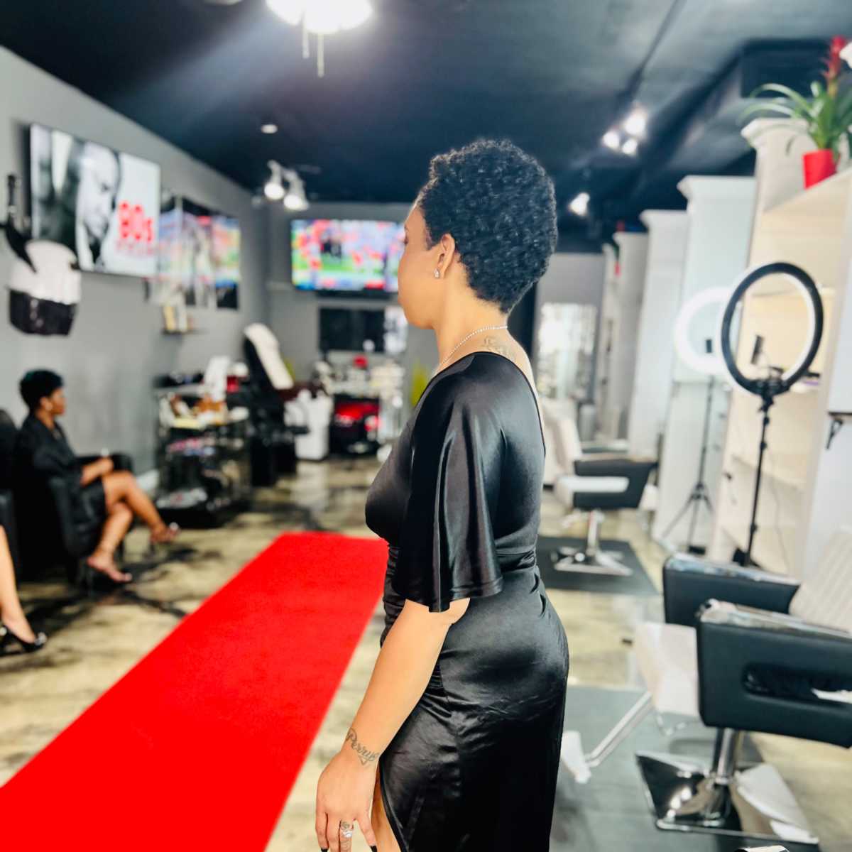 A woman in a black dress stands on a red carpet in a hair salon, while another seated woman is visible in the background.