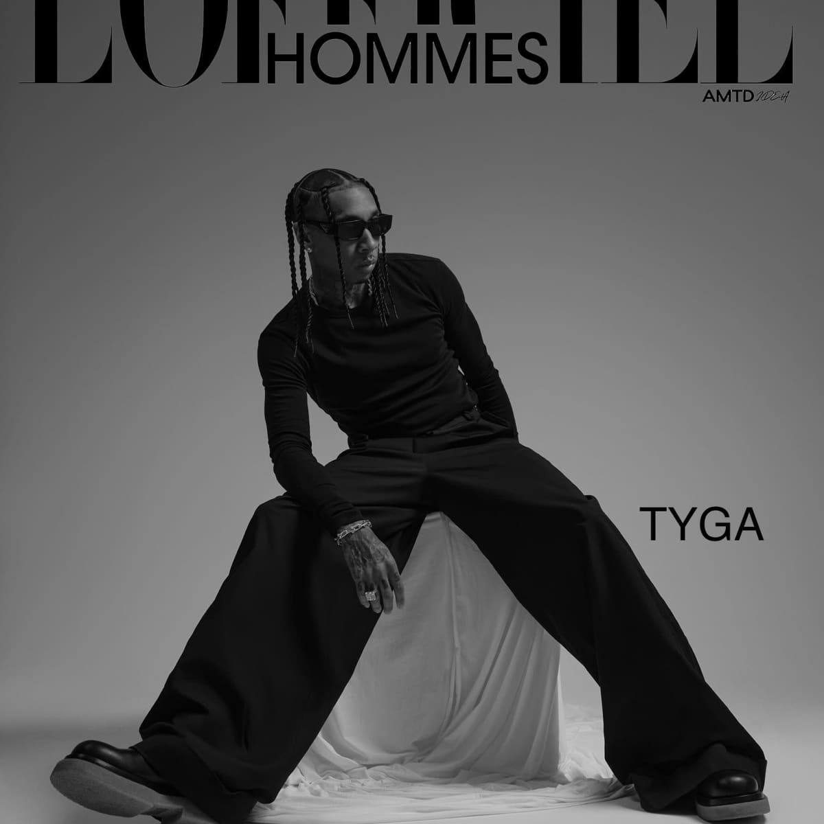 A person sitting on a white draped surface, dressed in a black outfit with wide pants and sunglasses, against a minimalistic background with the text "L'Officiel Hommes" and "TYGA."