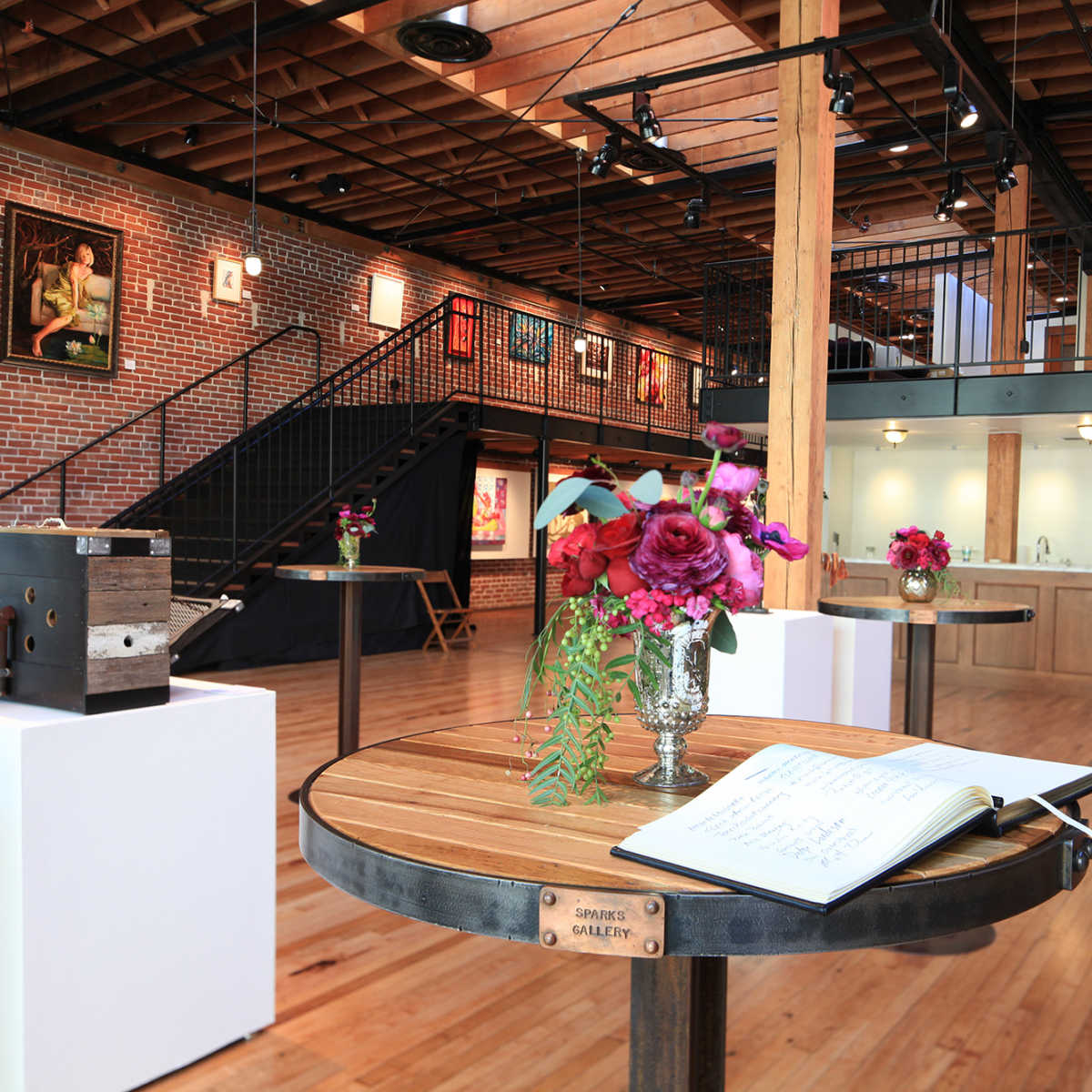 An art gallery interior with wooden floors, brick walls, tables with floral arrangements, and framed artworks hanging on the walls.