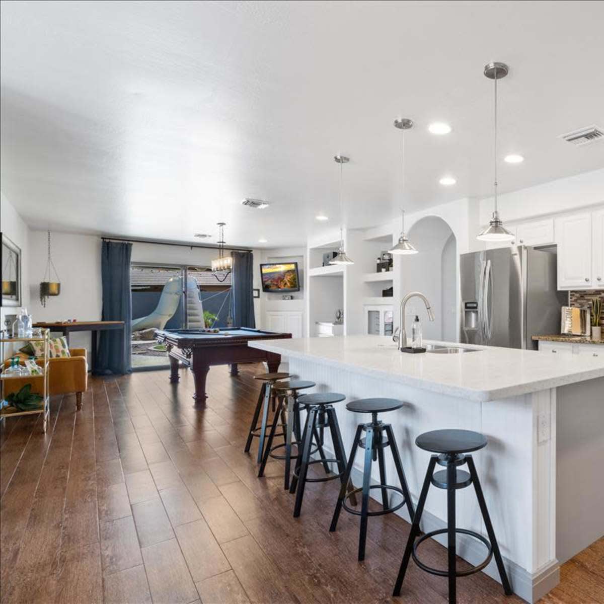 A modern kitchen with white cabinetry, a large island with bar stools, and a view into a spacious living area featuring a pool table.