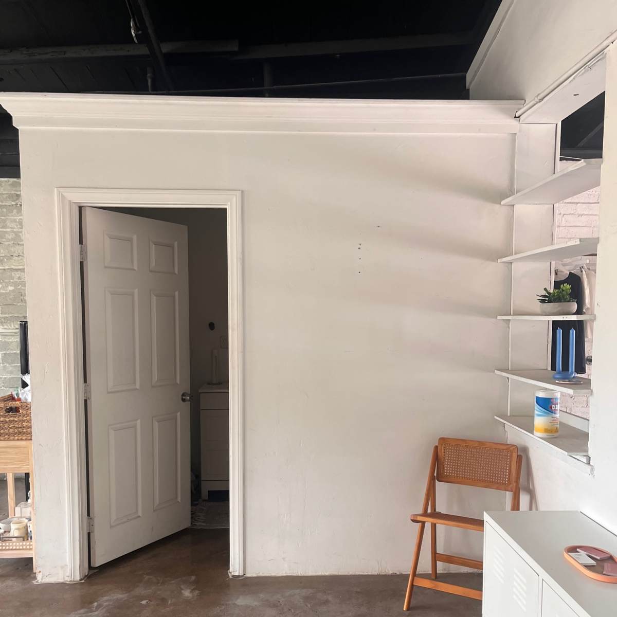 A small room features a white wall with a partially open door, a wooden chair, and a shelf with decorative items.