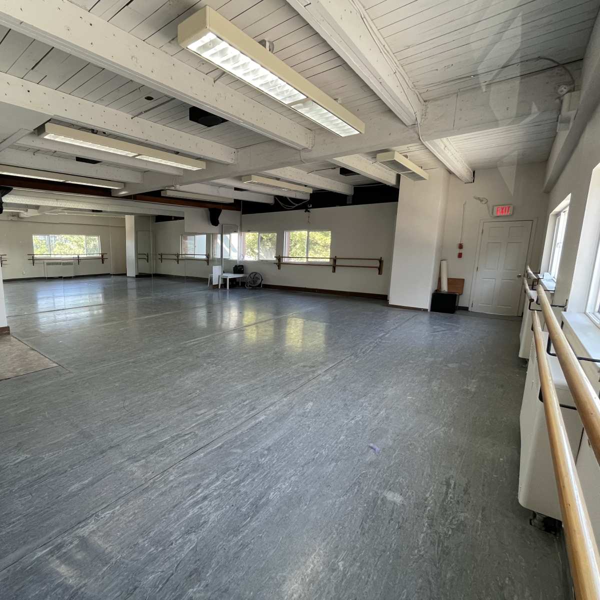 A spacious dance studio with wooden ballet barres along the walls, large windows allowing natural light, and a smooth flooring surface.