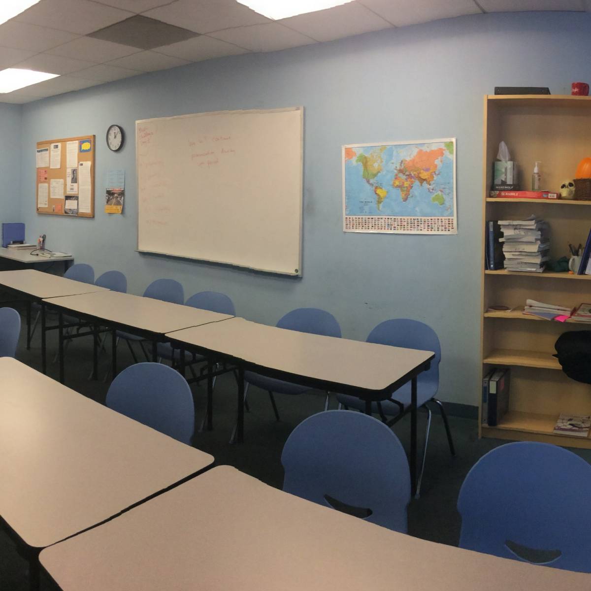 A classroom with rows of tables and chairs, a whiteboard, a map on the wall, and a bookshelf, is adjacent to a windowed area.