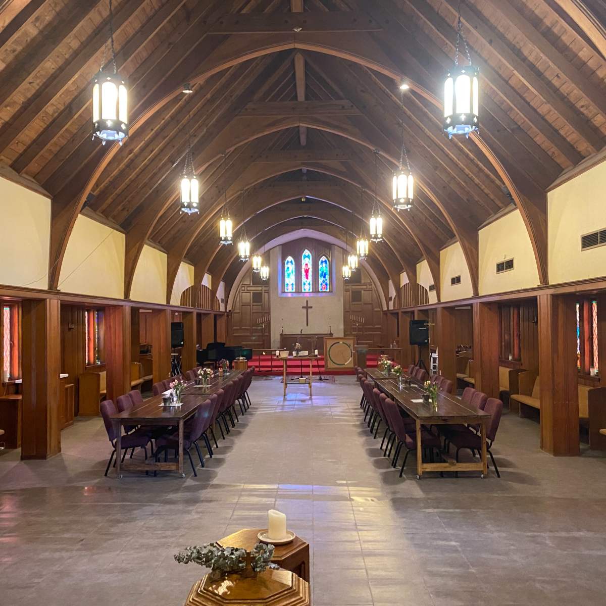 The interior of a church features wooden beams, stained glass windows, and rows of tables set for a gathering.
