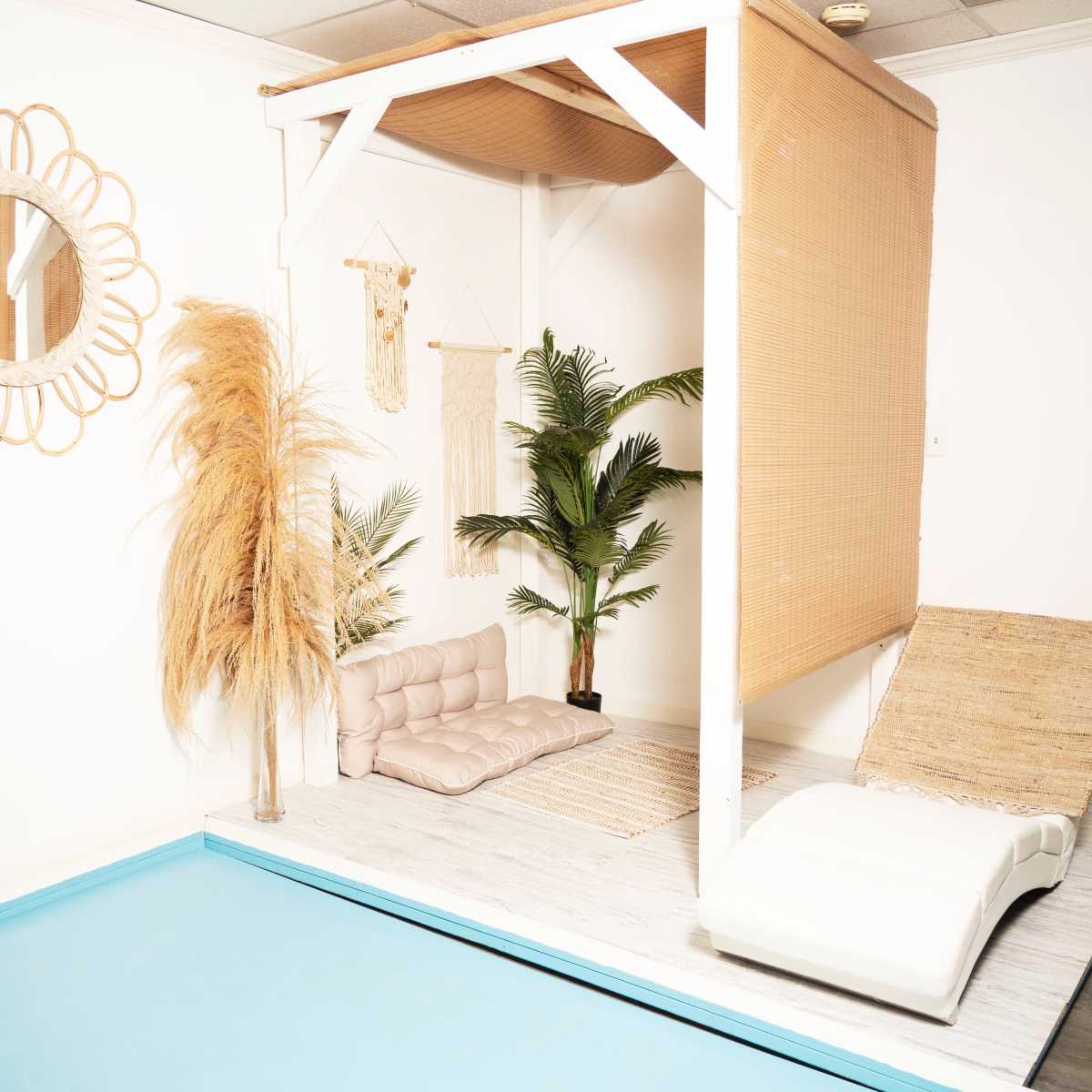 A relaxation area featuring a small pool, a raised platform with a cushion, indoor plants, and decorative wall hangings in a light-colored room.