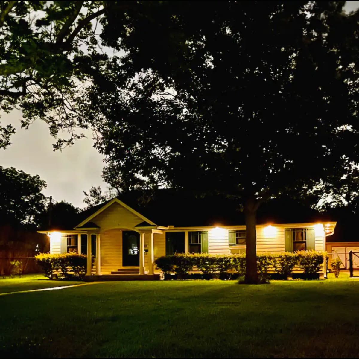 A single-story house with illuminated porch lights sits surrounded by trees in a dark, grassy yard at night.