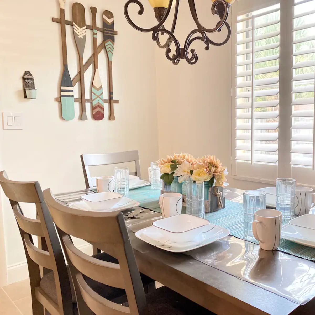 A dining table is set with plates, cups, and glasses, surrounded by chairs, with decorative paddles hanging on the wall and a flower arrangement in the center.